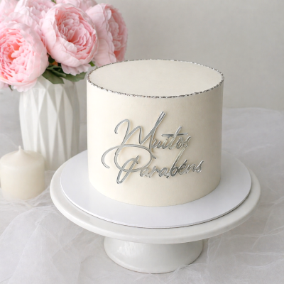 Bolo branco redondo com texto Muitos Parabéns em prateado, sobre prato branco, com flores cor de rosa e vela ao fundo.