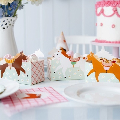 Caixas decorativas de papel com desenhos de cavalos sobre mesa branca com pratos e bolo decorado ao fundo