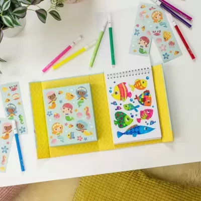 Caderno de desenho com sereias e canetas coloridas numa mesa branca