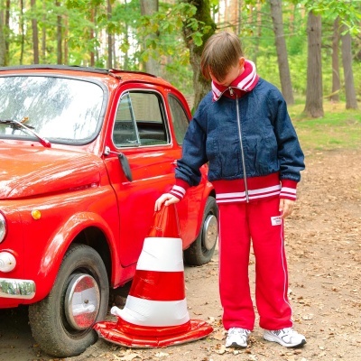Criança com roupa desportiva ao lado de carro vermelho e cone de sinalização