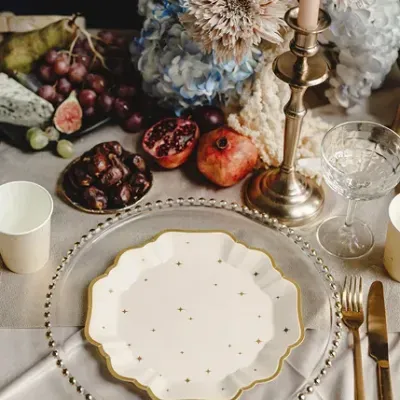 Mesa posta com pratos brancos e dourados, talheres dourados, vela, flores secas e frutas variadas