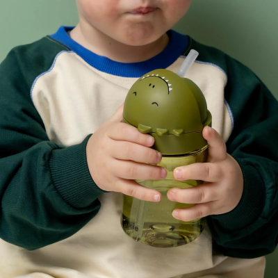 Copo infantil verde com tampa de dinossauro e canudo, mão de criança a segurar