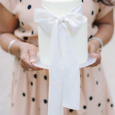 Bolo branco de duas camadas com laço decorativo segurado por pessoa com vestido bege de bolinhas pretas