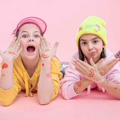 Duas raparigas deitadas com tatuagens coloridas nas mãos e braços, fundo rosa