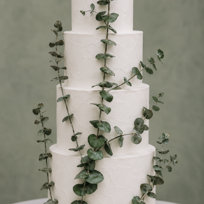 Bolo branco de quatro andares decorado com ramos verdes, em base de madeira num fundo cinza esverdeado.