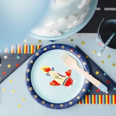 Conjunto de pratos de papel com desenhos de foguetes e estrelas, talheres plásticos, guardanapos e fitas coloridas sobre mesa azul
