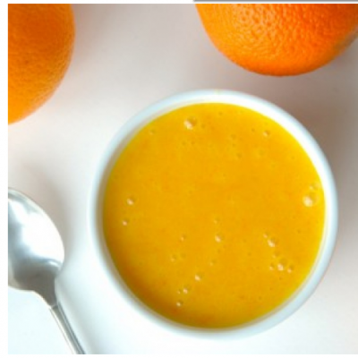 Tigela branca com líquido laranja cremoso, duas laranjas e colher metálica sobre fundo branco