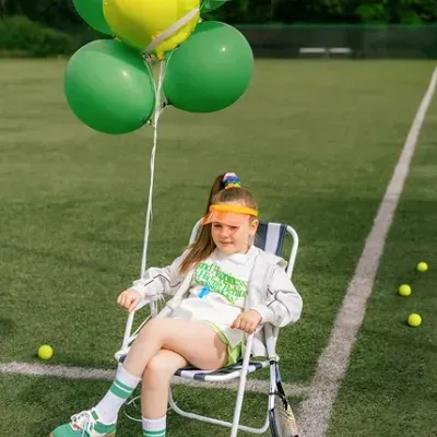 Menina sentada com roupas desportivas numa cadeira dobrável numa quadra de ténis com bolas e balões coloridos