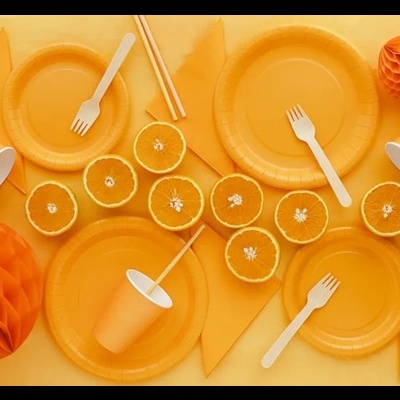 Artigos de festa laranja com pratos, copos, garfos, guardanapos, decorações em papel e laranjas cortadas ao meio