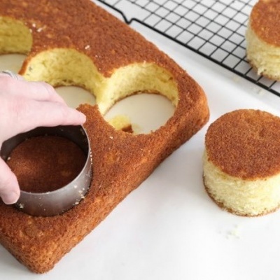 Bolo amarelo retangular com círculos cortados usando cortador metálico sobre superfície branca e grelha preta ao fundo