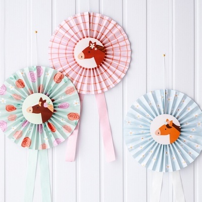 Rosetas de papel coloridas com desenhos de cavalos penduradas em parede branca de madeira