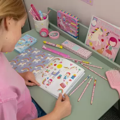 Mesa verde com material escolar com tema de unicórnios e criança a folhear livro de colorir.