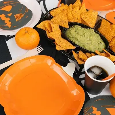 Pratos de papel laranja e cinzento com design de abóboras, copos pretos com canudos laranja, tortilhas amarelas com guacamole e tangerinas numa mesa com toalha preta e branca.