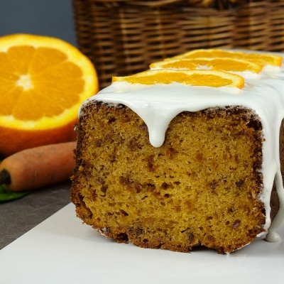 Bolo húmido com cobertura branca e fatias de laranja, cenoura e laranja ao fundo