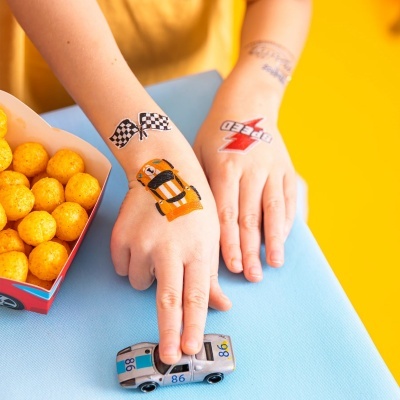 Duas mãos com tatuagens temporárias de corrida e um carro de brinquedo sobre uma mesa azul com um fundo amarelo