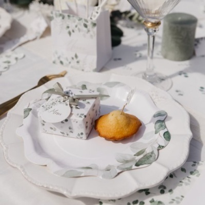 Pratos brancos com decoração verde, caixa de presente com etiqueta 'Merci', bolo e copo de vidro numa mesa decorada