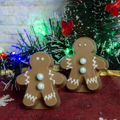 Dois bonecos de gengibre de chocolate com decoração de glacê branco e botões azuis em ambiente natalício com pinheiro artificial e luzes coloridas.