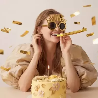 Mulher com óculos HAPPY BIRTHDAY dourados e bolo de aniversário amarelo com flores