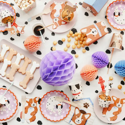 Artigos de festa decorados com tema de cães, incluindo pratos, copos e guardanapos com ilustrações de cães em tons naturais, bolas coloridas de papel e salgadinhos numa mesa branca.