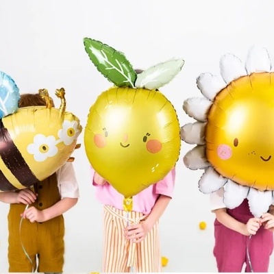 Três balões em forma de abelha, limão e girassol com crianças ao fundo