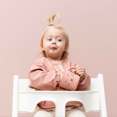 Bebé sentada em cadeirão branco segurando caneca rosa com rosto de gato.