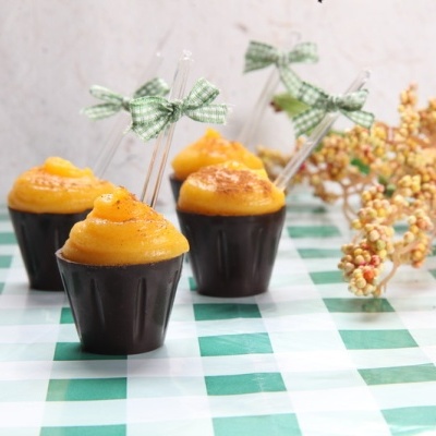 Copos pequenos com creme amarelo, colher com laço verde, sobre superfície xadrez verde e branca