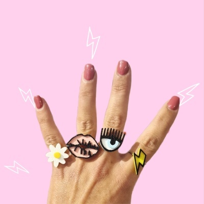 Mão com quatro anéis coloridos e unhas pintadas num fundo rosa