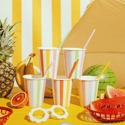 Cinco copos de papel com riscas coloridas e canudos rodeados por frutas e ambiente amarelo