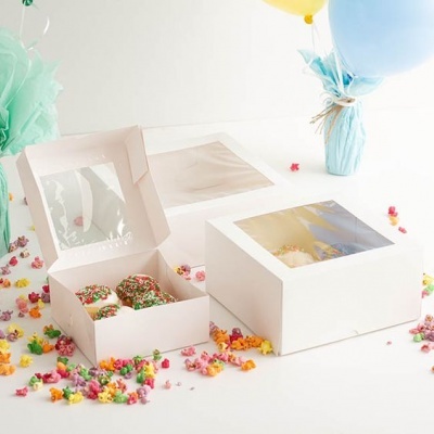 Caixas brancas de papel para bolos com janelas transparentes, bolos decorados e pipocas coloridas ao redor.