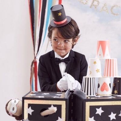 Criança com traje formal e chapéu pequeno a brincar com copos de papel empilhados em mesa decorada.