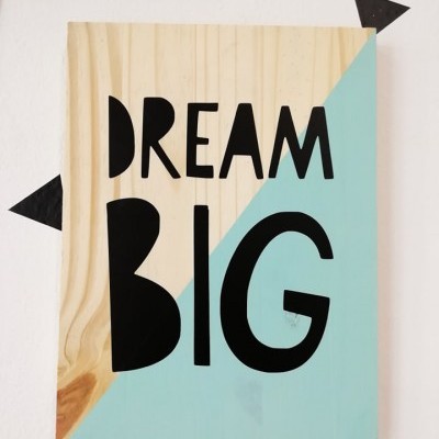 Placa em madeira natural e azul claro com texto preto DREAM BIG e triângulos pretos na parede