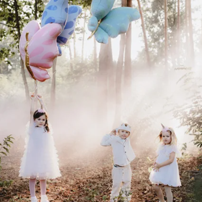 Crianças vestidas de branco segurando balões em forma de borboleta numa floresta