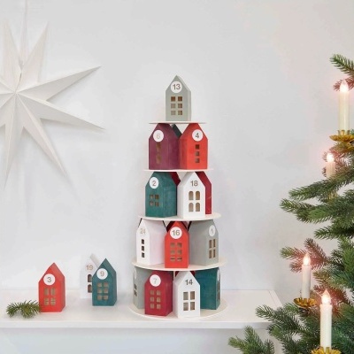 Casas de madeira decorativas em várias cores e números, empilhadas como árvore de Natal com velas e ramo de pinheiro ao lado