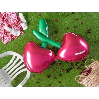 Bexigas metálicas em forma de cereja sobre relva com cadeira branca, sandálias, vestido floral e cesta de palha com cerejas