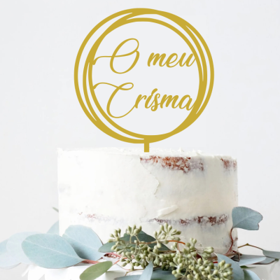 Bolo branco com decoração de folhas verdes e topo dourado com texto 'O meu Crisma'.