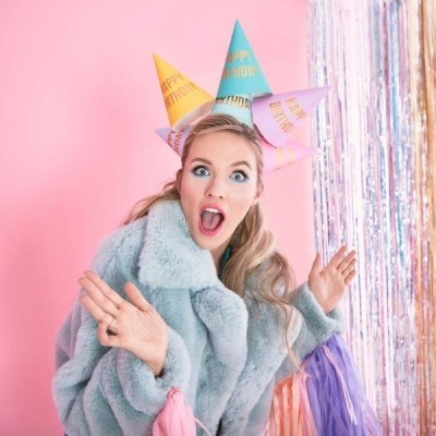 Mulher com casaco cinza e chapéus de festa coloridos em fundo rosa