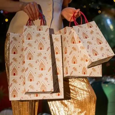 Três sacos de papel decorativos de Natal com padrão de gingerbread e biscoitos, segurados por pessoa com roupa dourada