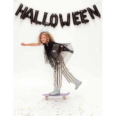 Menina em fato de Halloween com capa preta, calças às riscas, botas prateadas e coroa de aranha a andar de skate com balões a dizer HALLOWEEN