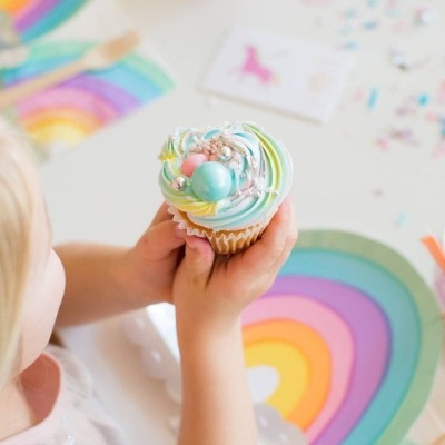 Criança segurando cupcake decorada com glacê colorido e decoração de arco-íris na mesa