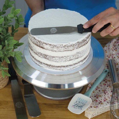 Bolo de camadas com cobertura branca em prato giratório de metal, espátula a alisar topo e utensílios de cozinha ao lado