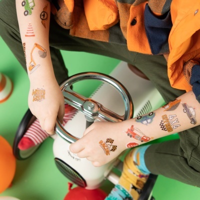 Criança com tatuagens temporárias de veículos e sinais de trânsito a segurar volante de carro de brincar branco.