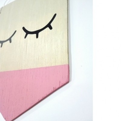 Placa decorativa em madeira com desenho de olhos fechados em preto e partes pintadas de creme e rosa