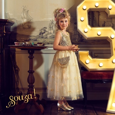 Menina vestida com vestido dourado e bege, sapatos dourados, bolsa pequena prateada, em ambiente interior clássico com mesa e luzes decorativas