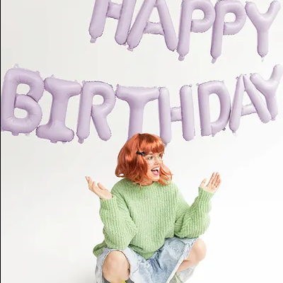 Balões lilás formando a frase HAPPY BIRTHDAY com mulher de suéter verde e botas amarelas frente a fundo branco