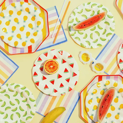 Pratos de papel com padrões de frutas, guardanapos coloridos, bananas, melancia e óculos de sol sobre mesa amarela