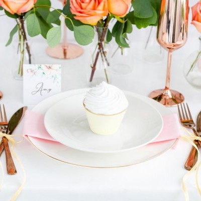 Mesa com prato branco, cupcake, talheres dourados, guardanapo rosa, copos rosé gold e flores laranja