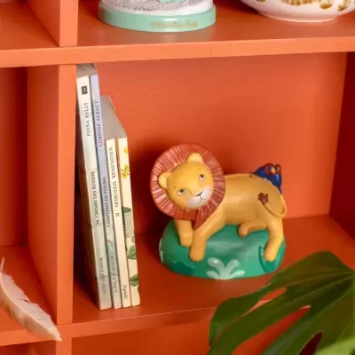 Estante cor de tijolo com livros, figura decorativa de leão amarelo com pássaro azul, pena branca e folha verde.