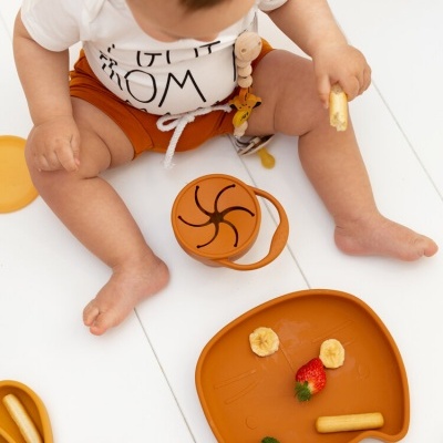 Bebé com prato e taza de silicone laranja com pedaços de fruta e legume