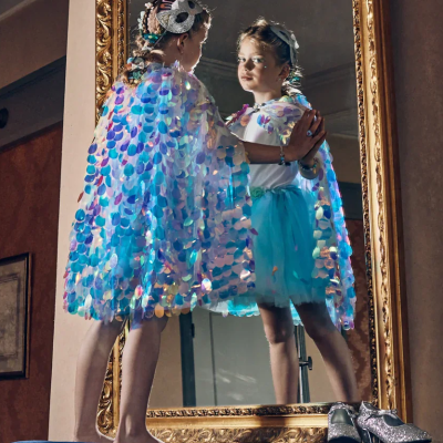 Menina com capa azul-turquesa de lantejoulas e saia de tule azul olhando para espelho dourado