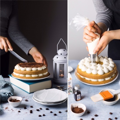 Bolo de camadas com chantilly e cacau em pó numa base giratória, lado de lanterna branca acesa e mãos a decorar com saco pasteleiro.